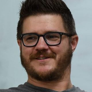 Portrait d'un homme barbu aux lunettes noires et t-shirt gris, nommé au Prix de l'Opticien International de l'Année 2025
